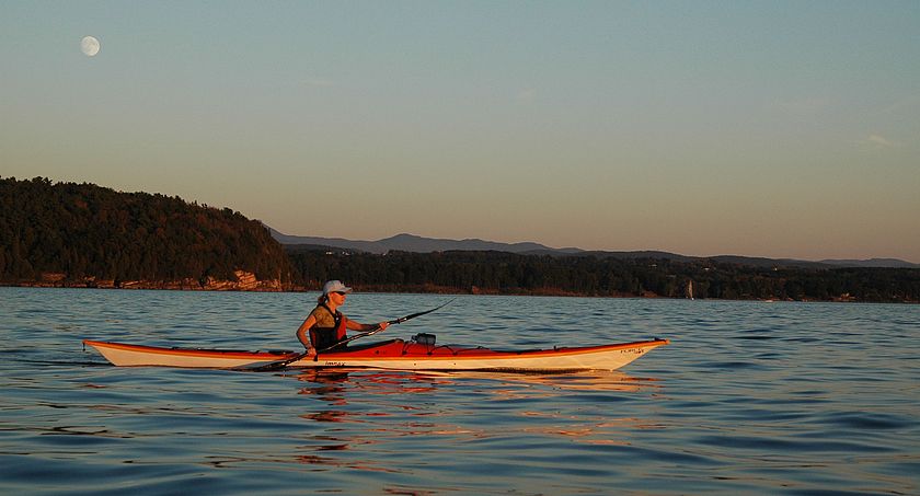 A paddler in a kayak on Lake Champlain at dusk with a full moon.