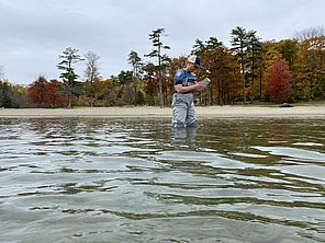 Photo of a woman in waders standing in a lake with autumn trees behind her on the shoreline.